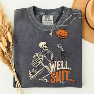 Peut inclure: Un t-shirt gris anthracite avec une illustration représentant un squelette assis tenant une ficelle reliée à une citrouille d'Halloween orange. Le texte "WELL, SHIT..." est imprimé en orange et crème sous le squelette. Col rond.