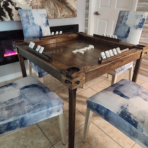 May include: A square wooden game table with a dark brown finish, featuring four corner cup holders and integrated racks for dominoes. The table is surrounded by four chairs upholstered in a blue and white abstract patterned fabric.
