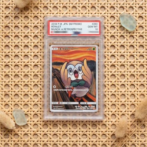 Puede incluir: Una carta coleccionable Pokemon Rowlet Munch: A Retrospective de 2018, graduada y sellada en una funda de plástico transparente. La carta muestra un búho de dibujos animados con las alas en la cara, imitando El Grito, con fondo naranja y azul.