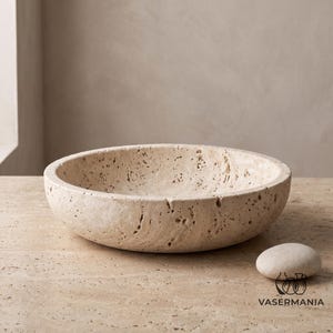 May include: A round, beige travertine stone vessel sink with a porous, textured surface. The basin features natural pits and variations in the stone, showcasing a circular shape with thick, sturdy walls. A small, smooth, oval stone sits nearby.