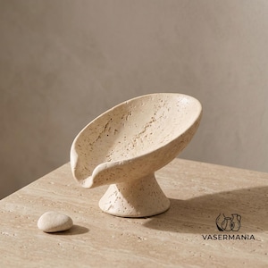 May include: A beige travertine soap dish with a porous, textured surface and a pedestal base, featuring a curved, shell-like bowl with a small spout. A single small, smooth, rounded beige stone sits beside the soap dish. Text reads VASERMANIA.