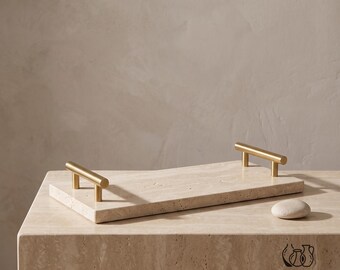 May include: A rectangular beige travertine tray with a porous, natural stone texture. The tray features two brushed gold-tone metal handles attached to the short ends. A small, smooth, rounded beige stone sits on the surface near the tray.