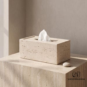 May include: A rectangular beige travertine tissue box cover with a porous, natural stone texture. A single white tissue protrudes from the central oval opening. The box features a clean, block-like construction with visible surface pitting.