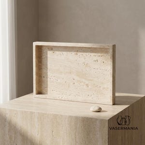May include: A rectangular beige travertine tray with a natural porous texture and visible stone veining. The tray features a raised edge and a smooth, flat interior surface. A small, smooth, rounded stone pebble rests on the surface next to the tray.