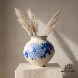 Puede incluir: Un jarrón de cerámica de color crema con forma redonda y superficie texturizada, pintado a mano con motivos florales azules. El jarrón contiene tres tallos de hierba de la pampa beige, altos y esponjosos. El logo VASERMANIA aparece abajo a la derecha.
