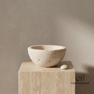 Peut inclure: Un bol en travertin beige avec une surface poreuse et texturée et une forme arrondie. Un petit galet en pierre lisse assorti est posé à côté du bol sur un piédestal carré en travertin. Le texte VASERMANIA est imprimé dans le coin inférieur droit.