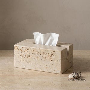 May include: A rectangular tissue box cover crafted from beige travertine stone with a natural porous, pitted texture. The top features a central rectangular slot with a white tissue protruding from it. The logo VASERMANIA is printed in the corner.