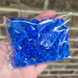 May include: A clear plastic bag filled with numerous small, faceted, translucent blue acrylic gems. Each individual piece has a geometric, diamond-like shape with multiple flat surfaces that catch the light, creating a bright, vibrant blue appearance.