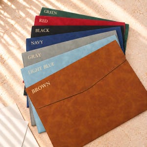 May include: A stack of rectangular document folders in various colours including brown, light blue, grey, navy, black, red, and green. Each folder features a flap closure and is labelled with its respective colour name printed in gold lettering.