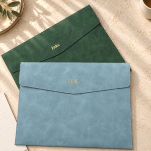 Puede incluir: Una carpeta de documentos de color verde oscuro con el nombre Jake grabado en oro y una carpeta de color azul claro con las iniciales EFK grabadas en oro. Ambas carpetas tienen un acabado mate liso y un diseño de solapa puntiaguda.