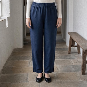 May include: A pair of solid navy blue trousers featuring an elasticated waistband and a straight-leg cut. The trousers are worn with a long-sleeved white top and simple black flat shoes, creating a clean and functional look for everyday wear.