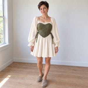 May include: A cream-colored mini dress featuring long, puffed sleeves with ruffled cuffs and a square neckline. The dress is layered with an olive green, heart-shaped corset bodice that has side ties, creating a structured silhouette.