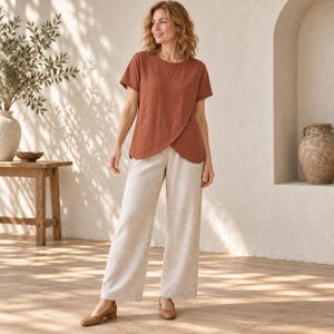 Op de afbeelding: Een roestkleurige top met korte mouwen en een afgeronde, overlappende zoom, gecombineerd met een wijde broek in een lichte beige neutrale tint. De top heeft een textuur in de stof, terwijl de broek een recht model en volledige lengte heeft.