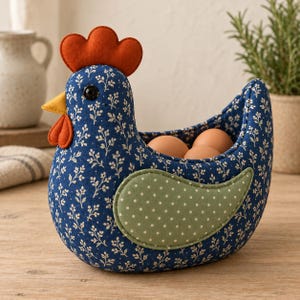Puede incluir: Una cesta de tela con forma de gallina que presenta un cuerpo azul oscuro con un patrón floral blanco, una cresta y barbillas de fieltro naranja brillante, un pico amarillo y un ala verde salvia con lunares blancos, conteniendo tres huevos marrones.