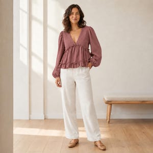 May include: A mauve long-sleeve blouse with a deep V-neckline, gathered waist, and ruffled hem detail paired with off-white wide-leg trousers. The blouse features full sleeves with cuffs, and the trousers have a straight-leg cut and neutral tone.
