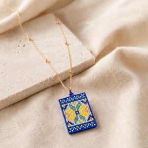 May include: A gold chain necklace featuring a rectangular pendant crafted from small blue, yellow, white, and green beads. The pendant displays a geometric floral pattern. The delicate gold chain includes small gold beads spaced along its length.