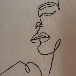 Puede incluir: Una pieza de arte mural que presenta un dibujo de una sola línea negra continua de un perfil de rostro humano sobre un fondo beige claro texturizado. La obra muestra el contorno de la frente, la nariz, los labios y la barbilla, con una línea curva para el hombro.