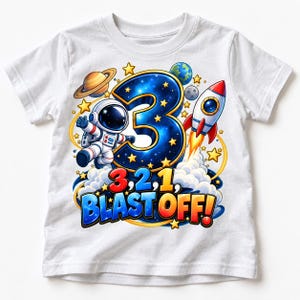 Könnte beinhalten: Ein weißes T-Shirt mit einem Weltraum-Motiv, das eine große blaue Zahl 3, einen Astronauten, eine Rakete, Planeten und Sterne zeigt. Der Text lautet 3, 2, 1, BLAST OFF! in bunten, fetten Buchstaben vor einem Hintergrund aus Wolken und Weltraum.