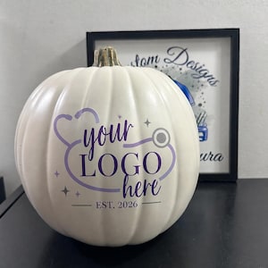 May include: A white artificial pumpkin featuring a purple graphic of a stethoscope forming a heart shape around the text "your LOGO here" and "EST. 2026" with small star accents. The pumpkin has a textured surface and a realistic stem on top.