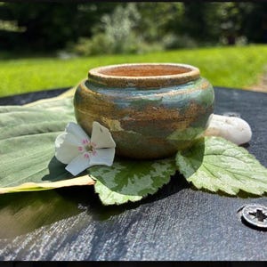 Peut inclure: Un petit bol en céramique fait main, de forme ronde, présentant une glaçure brillante dans des tons vert sauge et brun terreux. Le bol possède une large ouverture avec un bord épais et un corps arrondi, posé parmi des feuilles et une fleur blanche.