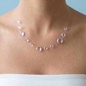Può includere: Una collana in stile invisibile caratterizzata da molteplici perline rotonde e trasparenti di varie dimensioni, distanziate lungo un sottile cordino di nylon traslucido. Le perline sembrano fluttuare attorno al collo.
