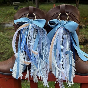 Puede incluir: Un par de botas vaqueras de cuero marrón adornadas con dijes decorativos. Cada dije presenta un gran lazo de tela azul claro sujeto a un anillo dorado, con cintas en cascada en tonos azul, blanco y beige, encaje y cuentas blancas.