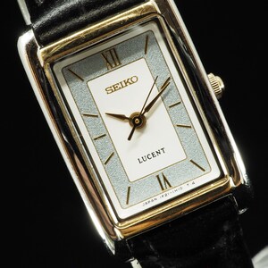 Puede incluir: Un reloj de pulsera rectangular Seiko Lucent con caja dorada, esfera blanca con borde plateado texturizado, manecillas doradas y números romanos en las posiciones de las doce y las seis, con una correa de cuero negro.