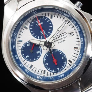 May include: A stainless steel Seiko Chronograph 100M watch with a white dial, three dark blue sub-dials, and red hands. The watch features a tachymeter scale on the blue outer bezel, a date window at the three o'clock position, and luminous hands.
