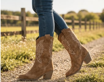 May include: Mid-calf tan suede boots featuring a stacked block heel and intricate white stitched embroidery patterns on the upper shaft. The boots have a rounded toe design and pull tabs at the top, worn with blue denim jeans.