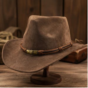 May include: A brown suede fedora hat featuring a pinched crown and a wide brim. The hat is accented with a decorative band made of thin brown leather cords and a central rectangular brass-toned metal plate embossed with a star-like emblem.