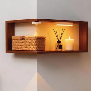 May include: A brown wooden corner shelf mounted on a wall, featuring integrated warm lighting. On the shelf sits a rectangular woven wicker basket, a glass reed diffuser with black sticks, and a white cylindrical pillar candle.