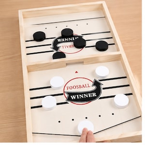 May include: A wooden tabletop game board featuring two divided sections with black and white circular pucks. The board surface displays black horizontal lines, red circular accents, and the text "FOOSBALL WINNER" printed in black and red.