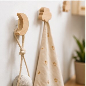May include: A set of light-colored wooden wall hooks featuring a crescent moon shape and a cloud shape. The moon hook holds a beige fabric item with a knotted rope loop, while the cloud hook supports a beige cloth with a small brown pattern.