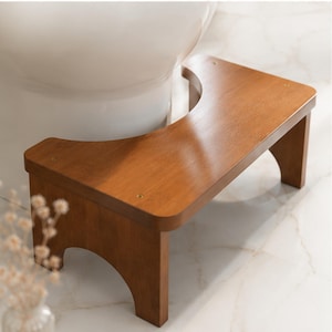 May include: A medium-brown wooden bathroom stool with a smooth, polished finish and a curved cutout along the back edge. The stool features four sturdy legs with arched openings and a flat, rectangular top surface designed to fit around a toilet base.