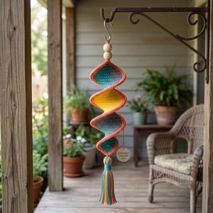 May include: A spiral-shaped crochet hanging ornament featuring alternating sections of orange, yellow, teal, and grey yarn. The piece includes two wooden beads at the top, a hanging hook, and a multi-colored tassel at the bottom with a round tag reading Crochet Valley 2024.