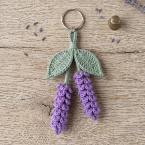 Puede incluir: Un llavero de ganchillo con dos tallos de flor de lavanda en hilo morado y dos hojas de color verde claro, unido a una anilla de metal plateado. Las flores de lavanda tienen textura con puntos pequeños y redondeados, y las hojas son planas.