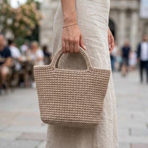 Op de afbeelding: Een beige gehaakte handtas met een gestructureerde trapeziumvorm en een enkele geïntegreerde handgreep. De tas is gemaakt van een dicht, uniform steekpatroon in een neutrale beige kleur, wat zorgt voor een getextureerd uiterlijk en open ontwerp.