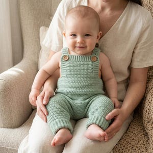 以下が含まれることがあります： セージグリーンのクロシェ編みベビー用ロンパース。スクエアネックで、足首にはリブ編みのカフス、ショルダーストラップには2つの小さな木製ボタンが付いています。全体に一貫した質感のある編み目模様が施されています。