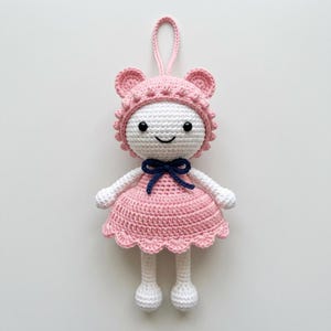May include: A handmade crochet doll featuring a white head with black eyes and a stitched smile, wearing a pink bonnet with small bobble details and a matching pink dress with a scalloped hem. A dark blue bow sits at the neck, and a loop is attached.