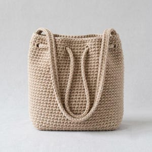 Puede incluir: Un bolso tipo cubo de color beige confeccionado con un patrón de tejido de ganchillo denso. El bolso presenta un cuerpo cilíndrico estructurado, una abertura ancha con ojales integrados y dos correas de ganchillo a juego.