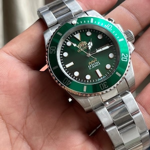 May include: A stainless steel wristwatch with a green dial and a matching green rotating bezel. The watch face features luminous hour markers, silver hands, and the text Orient, AAA Crystal, and 21 Jewels printed in white and gold lettering.