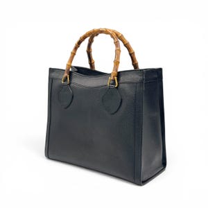 May include: A structured black leather handbag featuring a distinctive arched bamboo handle. The bag has a rectangular silhouette with gold-toned metal hardware connecting the handle to the main body, which displays a textured grain finish.