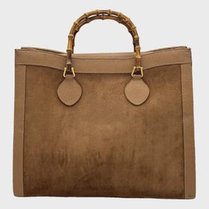 May include: A structured brown suede handbag featuring a distinctive arched bamboo handle. The bag has a rectangular body with smooth leather trim along the sides and top, complemented by gold-toned metal hardware attachments for the handle.