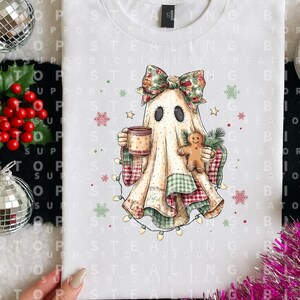 Peut inclure: Un t-shirt blanc avec un motif représentant un fantôme portant un nœud floral, tenant une tasse et un biscuit en pain d'épice. Le fantôme est enveloppé dans une couverture en patchwork aux motifs verts et rouges, entouré de flocons.