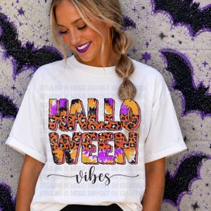 May include: A white short-sleeved t-shirt featuring the text HALLOWEEN in a bold, orange and purple leopard print pattern, with the word vibes written in a black cursive script underneath. The shirt has a crew neckline and cuffed sleeves.