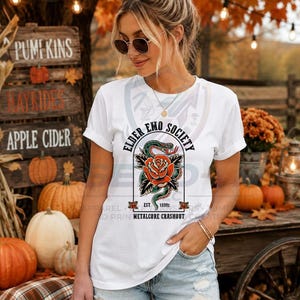 May include: A white short-sleeved t-shirt featuring a graphic print of a coiled green snake around a large orange rose. The text reads ELDER EMO SOCIETY, EST 1990s, and METALCORE CRASHOUT. The shirt has a crew neckline and rolled sleeve cuffs.