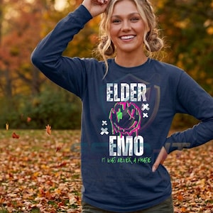 May include: A navy blue long-sleeve shirt featuring a graphic print with the text ELDER EMO and IT WAS NEVER A PHASE. The design includes a distressed white font and a melting smiley face graphic in neon pink and green with X-shaped eyes.