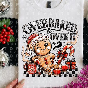 May include: A white t-shirt featuring a graphic of a winking gingerbread person wearing a Santa hat. The text reads OVERBAKED & OVER IT. The design includes a red mug with a snowflake, a candy cane, a gift box, and a black and white checkered base.