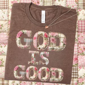 May include: A brown short-sleeved t-shirt featuring the text "GOD IS GOOD" in large, fabric-appliqué letters. The letters are crafted from various patches of floral, plaid, and gingham fabric in shades of pink, white, and brown. A thin necklace is visible.