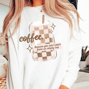 May include: A white long-sleeve sweatshirt featuring a graphic of a glass with a brown and white checkered pattern. The text reads coffee... because you can't pour from an empty cup. The design includes small star accents and a straw in the glass.